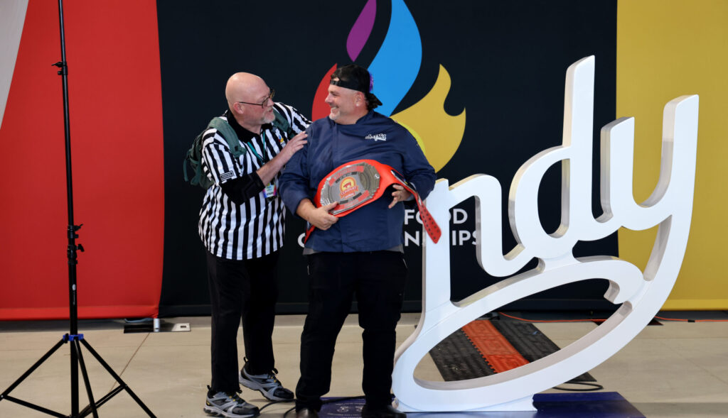 Chef Chefski celebrates at the World Food Championships in Indianapolis, holding the competition belt on stage with an event official beside the large “Indy” display sign.