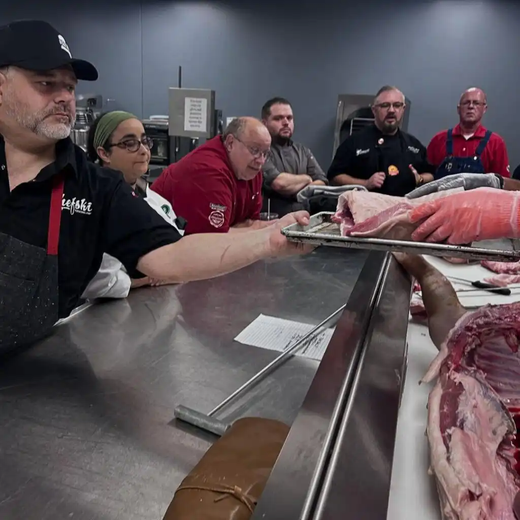 Two men in red chef uniforms handing a tray of fresh raw pork loin across a stainless steel butcher table in a professional kitchen