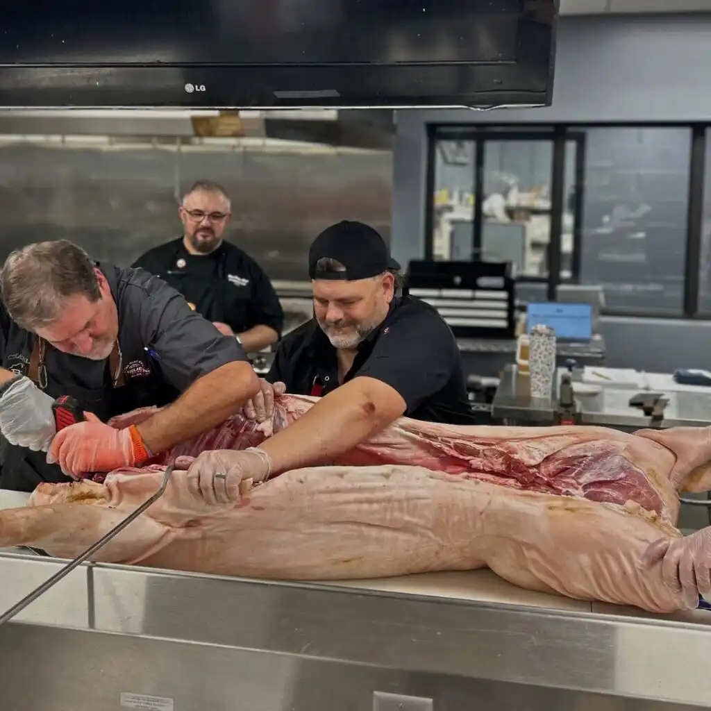 Chefs breaking down a whole pig on a stainless steel table in a commercial kitchen